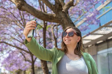 Gülümseyen iş kadını, şehrin ortasında çiçek açan jakaranda ağaçlarıyla akıllı telefonuyla selfie çekiyor.
