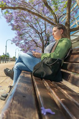 Yeşil ceketli bir kadının güneşli bir parktaki çiçek açan jakaranda ağaçlarının altında otururken deftere yazdığı dikey bir fotoğraf..