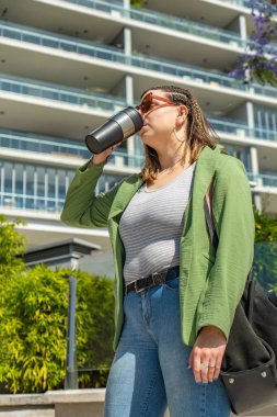 Güneşli bir günde modern bir binanın önünde kahvesini yudumlayan bir kadın.