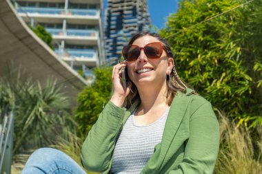 Genç iş kadını yeşil bitkiler ve binalarla dolu modern bir şehir ortamında cep telefonuyla konuşurken gülümsüyor.