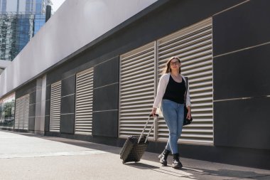Genç bir iş kadını modern bir binanın yanındaki kaldırımda yürüyor, bavul çekiyor ve omuz çantası taşıyor, havaalanına ya da tren istasyonuna iş gezisine gidiyor.