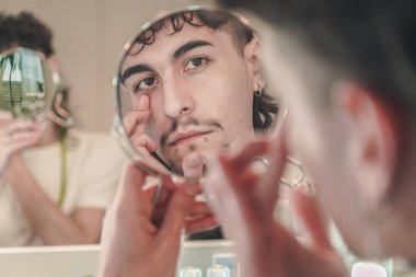 Piercingli ve şekilli saçlı genç adam, kendi algısı, kimliği ve çeşitliliği modern bir banyo ortamında keşfeden, küçük bir el kamerası aynasında yüzünü inceliyor.