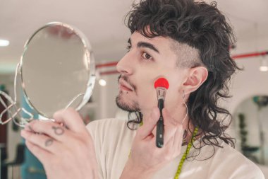 Dövmeleri ve piercingleri olan genç bir adam, parlak ışıklı bir odada küçük bir ayna tutarken, elmacık kemiklerine allık sürerek, erkek güzelliğine ve özgüvenine modern bir yaklaşım sergiliyor.