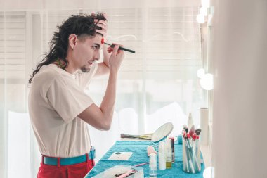 Uzun, kıvırcık saçlı, alnına makyaj yapan genç bir adam. Parlak bir ayna önünde, kendini ifade eden ve güzellik rutinlerinde çeşitlilik gösteren.