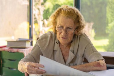 Gözlük takan yaşlı bir kadın, ofis masasında otururken önemli belgeler okuyor, evrak işlerine ve idari görevlere konsantre oluyor.