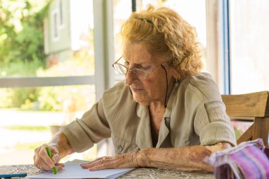 Gündüz vakti pencerenin yanındaki ahşap masada otururken yeşil renkli kalemle çizim yapan konsantre yaşlı kadın.
