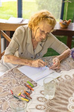 Kıdemli bir kadın, bir bardak suyla bir masada pastel boyalarla resim çizerek, yaratıcılığını ifade ederek ve rahatlatıcı bir anın tadını çıkararak boş zamanlarının tadını çıkarıyor.