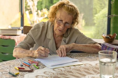 Gözlüklü yaşlı bir kadın bir bardak suyla bir masada pastel boyayla resim çizmeye odaklanmış. Parlak, rahat bir ev ortamında rahatlatıcı bir hobiden keyif alıyor.