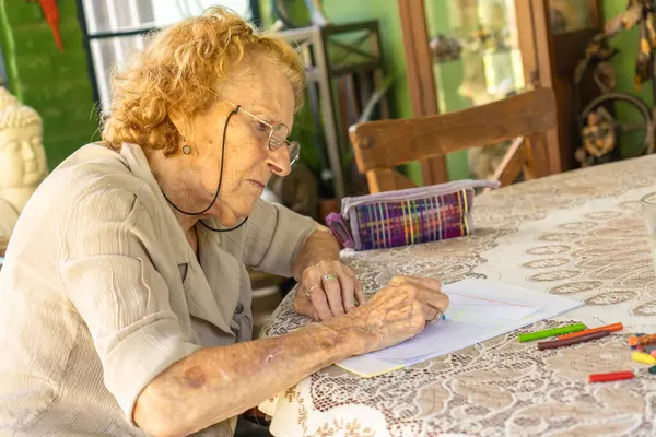 Yaşlı bir kadın kendini pastel boyalarla çizime kaptırmış bir şekilde masada oturuyor, evde yaratıcı aktiviteler yaparak tatmin edici bir emeklilik sergiliyor.