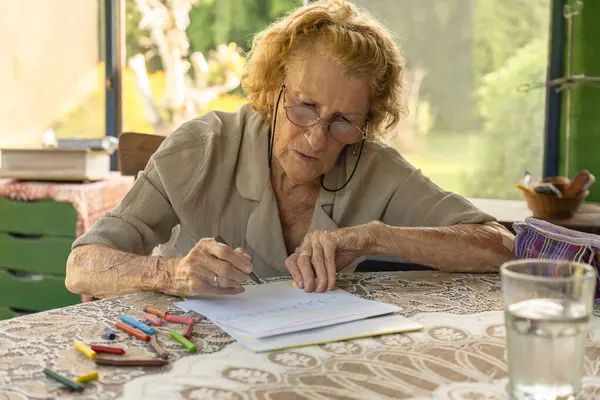 Gözlüklü yaşlı bir kadın bir bardak suyla bir masada pastel boyayla resim çizmeye odaklanmış. Parlak, rahat bir ev ortamında rahatlatıcı bir hobiden keyif alıyor.