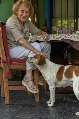 Üst düzey bir kadın terasta ahşap bir masada rahatça oturuyor, melez köpeğini nazikçe okşuyor, yazın sıcaklığında huzurlu bir bağ ve şefkat anının tadını çıkarıyor.