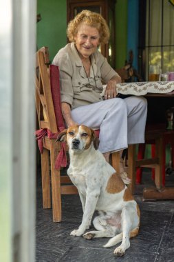 Renkli evinde bir masada oturan yaşlı kadın, melez köpeğini nazikçe okşuyor, sevgi ve arkadaşlıkla dolu huzurlu bir anı paylaşıyor, mutluluk ve rahatlık yayıyor.