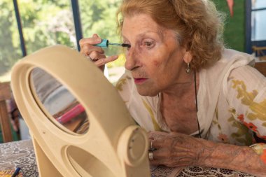 Masada küçük bir ayna kullanarak rimel süren yaşlı bir kadın, güzellik rutinini hassasiyetle ve dikkatle geliştiriyor, özgüven ve özgüveni somutlaştırıyor.