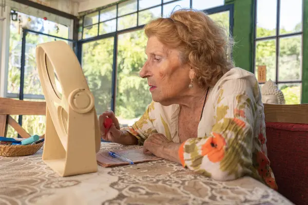 Üst düzey bir kadın evdeki bir masaya yerleştirilmiş küçük bir aynaya bakarken ruj sürüyor. Pencereden gelen doğal ışık rahat ve rahat bir atmosfer yaratıyor.