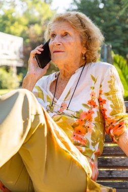 Park bankında rahat bir şekilde oturan kıdemli kadın, bir telefon görüşmesi yapıyor ve sıcak yaz havasının tadını çıkarırken aynı zamanda sevdikleriyle sakin bir açık hava ortamında iletişim kuruyor.