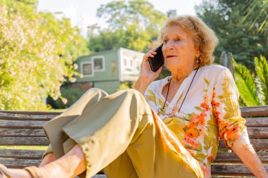 Huzurlu bir bahçe ortamında, ahşap bir bankta, yeşil ışıklarla çevrili ve sıcak güneş ışığıyla yıkanmış bir şekilde konuşurken, cep telefonuyla sohbet eden yaşlı bir kadın.