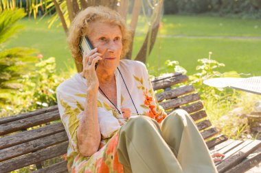 Üst düzey bir kadın, sakin bir park ortamında ahşap bir bankta oturmuş, teknolojiyi kucaklıyor ve emekliliğine bağlı kalarak cep telefonuyla konuşmaktan zevk alıyor.