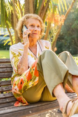 Kıdemli kadın, yemyeşil yemyeşil bir bankta otururken ve sıcak güneş ışığıyla yıkanırken akıllı telefonuyla konuşmaktan zevk alıyor.