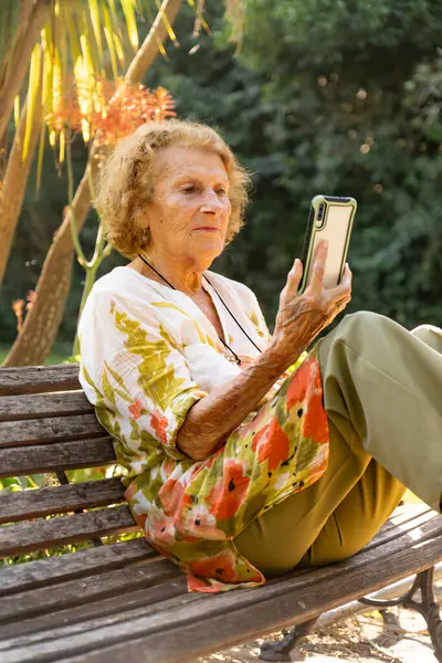 Yaşlı kadın parkta huzurlu bir anın tadını çıkarıyor, akıllı telefonunu karıştırırken rahat bir şekilde bankta oturuyor ve sıcak güneş ışığı altında etrafındaki dünyayla bağlantı kuruyor.