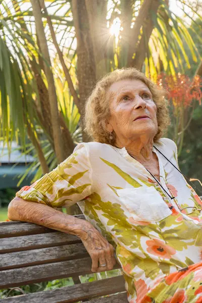 Kıdemli kadın yemyeşil bir bahçede huzurlu bir anın tadını çıkarıyor, ahşap bir bankta oturuyor güneş ışığı titreyen yaprakların arasından yavaşça süzülüyor, doğanın huzurunu kucaklıyor.