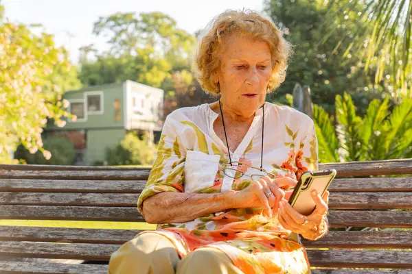 Sunny Park 'ta ahşap bir bankta oturan yaşlı kadın, sıcak havanın ve doğanın güzelliğinin tadını çıkarırken bağlı kalmak için cep telefonu kullanıyor.