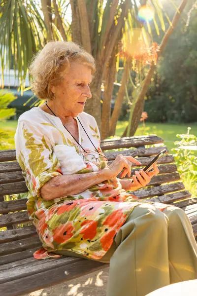 Park bankında oturan son sınıf öğrencisi kadın güneş ışığının tadını çıkarıyor ve akıllı telefonunda geziniyor. Teknoloji ve yaşlanma kavşağını gösteriyor.