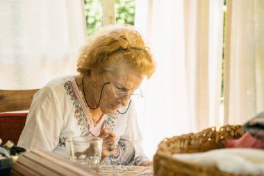 Yaşlı kadın, gözlük takıyor, iğnelerine konsantre oluyor, ustalıkla evinin huzurlu ortamında karmaşık bir nakış yapıyor.