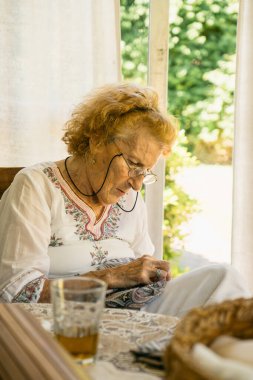 Pencerenin kenarında oturan yaşlı kadın, doğal ışıkla yıkanmış, kumaş dikiyor ve bir bardak çayın tadını çıkarıyor, huzurlu bir öğleden sonranın sükunetini somutlaştırıyor.