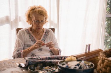 Konsantre üst düzey terzi titizlikle iğneyi iplikçikleyerek, evdeki bir masada dikiş malzemeleriyle çevrili, doğal ışıkla aydınlatılmış.