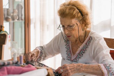 Kıdemli terzi kumaşı ev atölyesinde yoğunlaşmış, renkli iplikler ve dikiş aletleriyle çevrili, zanaatkar kendini adamışlık ve beceri örneği.