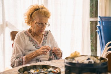 Üst düzey kadın kumaşı dikkatlice dikiyor, iğne ve iplik kullanıyor, düğme ve dikiş malzemeleriyle dolu bir masada, aydınlık, ev ortamında rahatlatıcı bir hobi ediniyor.