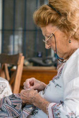 Odaklanmış yaşlı terzi titizlikle kumaşı iğne ve iplikle dikiyor. Parlak, rahat bir ortamda terziliğe karşı uzmanlık ve tutku gösteriyor.
