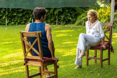 Kıdemli kadın ve fizyoterapisti bahçede sandalyelerde oturup hareket ve refahı artırmak için rehabilitasyon egzersizleri yapıyorlar.