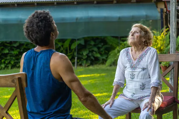 Üst düzey kadın ve fizyoterapisti bir bahçede sandalyelerde oturup birlikte nefes egzersizleri yapıyor, solunum sağlığı ve refahı geliştirmeye odaklanıyor.