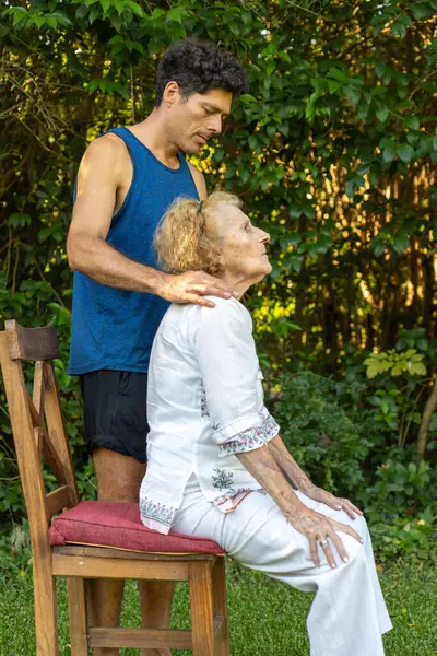 Fizyoterapist, kıdemli bir kadına duruş egzersizlerinde rehberlik ediyor, sakin bir açık hava ortamında, hareketliliği ve refahı teşvik eden bir sandalyede otururken destek ve nazik ayarlamalar sağlıyor.