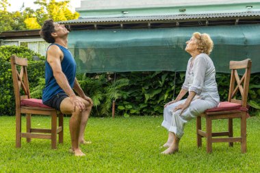 Üst düzey kadın ve kişisel antrenör, sakin bir bahçede sandalyeler üzerinde oturma egzersizleri yapıyor, sağlıklı yaşlanma ve açık hava fitness aktiviteleri yoluyla hareket kabiliyetini destekliyor.