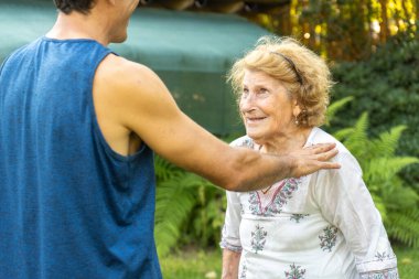 Genç fizyoterapist yaşlı bir kadını destekliyor ve rahatlatıyor. Rehabilitasyon sırasında bahçeye çıkıyor, yaşlılıkta sağlık ve refahı destekliyor.