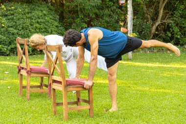 Son sınıf öğrencisi erkek ve kadın bahçede yoga yapıyor, denge pozisyonu yapıyor destek için sandalyeler kullanıyor, sağlıklı yaşam tarzını ve yaşlılığı destekliyor.