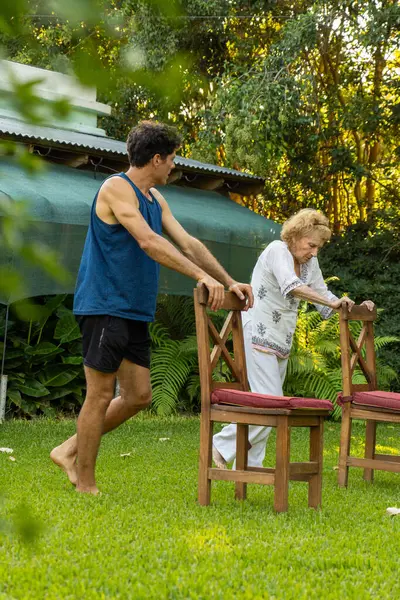 Genç erkek fizyoterapist yaşlı bir kadına rehabilitasyon egzersizlerinde yardımcı oluyor yemyeşil bir bahçede sandalyeler kullanıyor, hareketliliği ve iyileşmeyi teşvik ediyor.