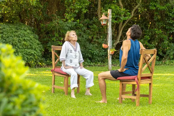 Üst düzey kadın ve fizyoterapisti bir bahçede sandalyelerde oturup boyun esnetme egzersizleri yapıyorlar, rehabilitasyon programının bir parçası olarak yukarı bakıyorlar.