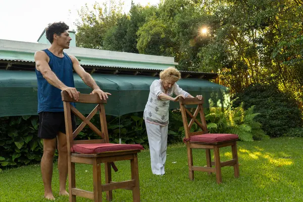 Kıdemli kadın kişisel antrenörünün yardımıyla fizik tedavi egzersizleri yapıyor, arka bahçesinde sandalyeleri destek olarak kullanıyor, yaşlılıkta sağlık ve hareketliliği teşvik ediyor.
