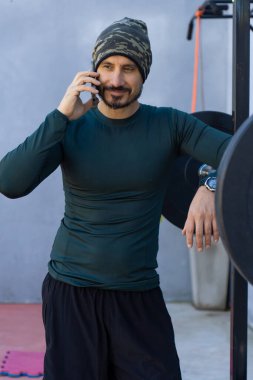 Sporcu antremanına ara verir, halter rafına yaslanır ve cep telefonuyla konuşur, fitness ve bağlantı karışımını gösterir.