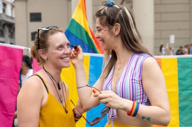 İki kadın gülümsüyor ve yüzlerine gökkuşağı makyajı yapıyor. Neşeli bir gurur geçidinde, çeşitliliği ve lgbtq + haklarını kutluyorlar. Renkli bayraklar ve neşeli bir atmosferle çevrili.