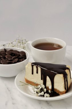 Koyu çikolatayla kaplı bir cheesecake. Bir fincan çay. Bir tabak çikolatalı fasulye. Beyaz çiçek 