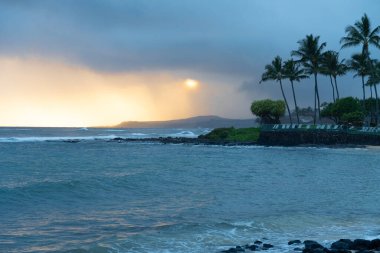 Hawaii, Kauai 'nin güney kıyısındaki Poipu Sahili' nde güzel bir akşam manzarası. Günbatımının ılık altın renkleri okyanusun sakin sularına yansıyor. Palmiye ağaçlarının silueti sönen ışığın yumuşak parıltısına karşı duruyor.