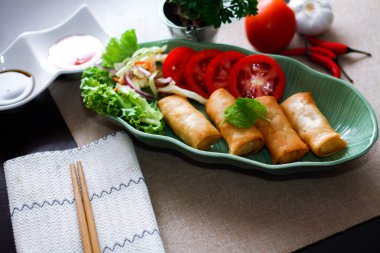 Kızarmış börekler, sebzeler ve domatesler yeşil yaprak şeklinde bir tabağa konmuş siyah ahşap bir masa ve Tayland sosu..