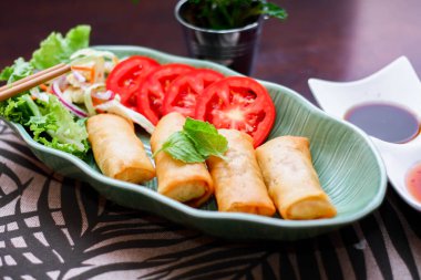 Kızarmış börekler, sebzeler ve domatesler yeşil yaprak şeklinde bir tabağa konmuş siyah ahşap bir masa ve Tayland sosu..
