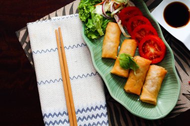 Kızarmış börekler, sebzeler ve domatesler yeşil yaprak şeklinde bir tabağa konmuş siyah ahşap bir masa ve Tayland sosu..
