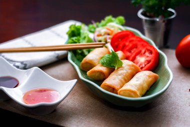 Kızarmış börekler, sebzeler ve domatesler yeşil yaprak şeklinde bir tabağa konmuş siyah ahşap bir masa ve Tayland sosu..