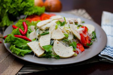 Gri bir tabakta servis edilen Vietnam sosisli Baharatlı Cam Erişte Salatası Ahşap masa ve çeşitli sebzeler.